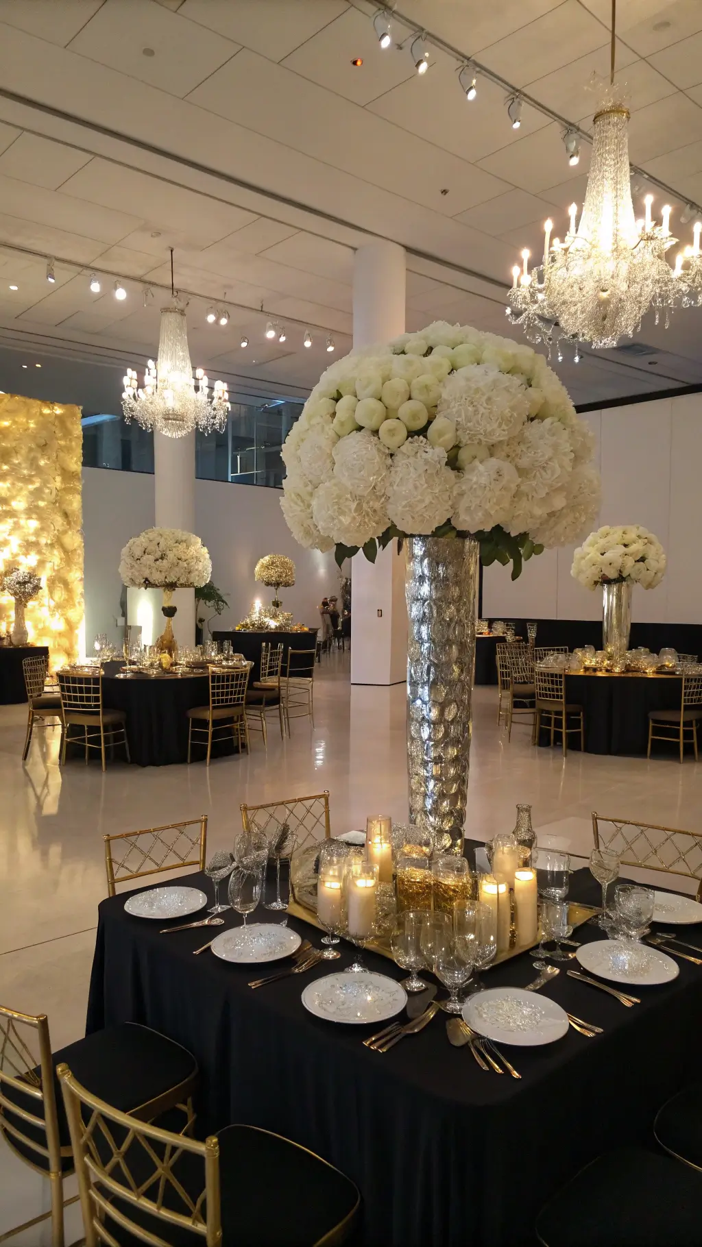 A luxurious reception in a modern art gallery features mirrored tables with black silk linens, adorned with tall arrangements of white roses, dahlias, and orchids in gold geometric vessels. Crystal candelabras and votives enhance the dramatic lighting, while ghost chairs with gold cushions complement the metallic accents. A massive floral wall installation serves as the focal point, and the low angle shot captures chandelier reflections on mirrored surfaces, creating an infinity effect.