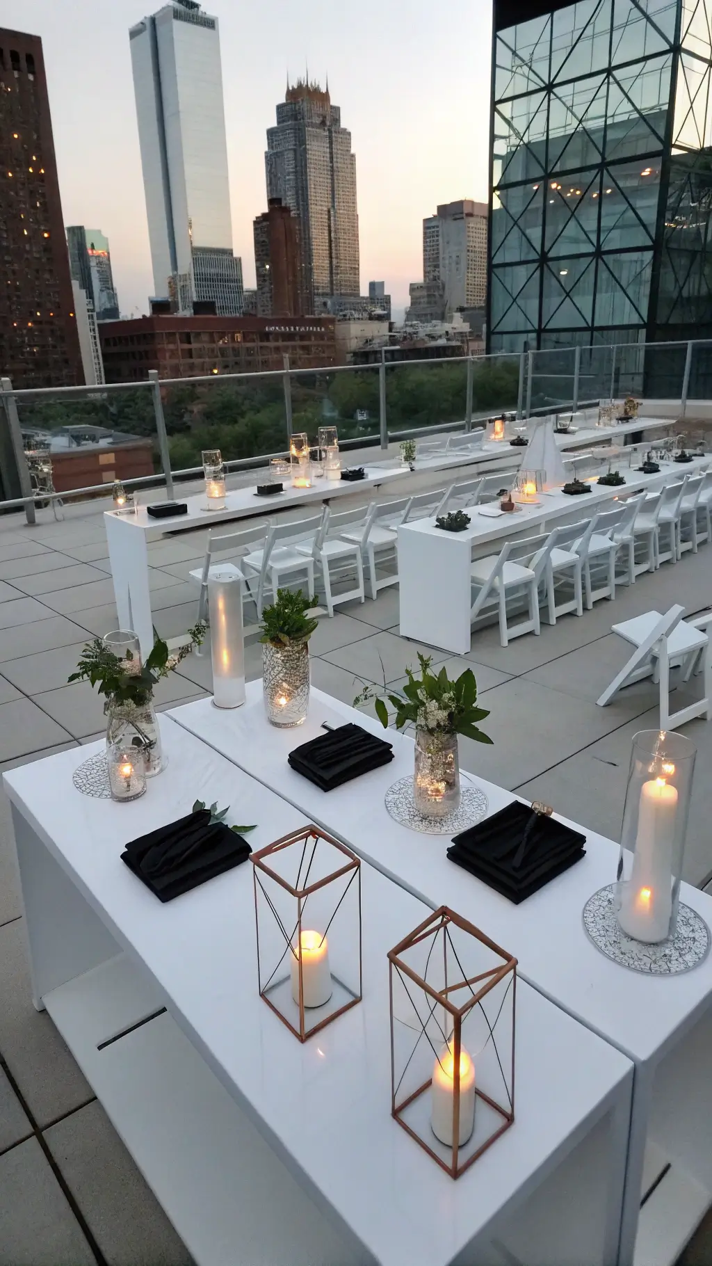 A rooftop ceremony at blue hour featuring modern minimalist decor with white acrylic tables, black matte chargers, dramatic calla lily stems in tall vases, and geometric copper terrariums with candles. Ghost chairs surround the setup, with city skyline visible through glass walls, creating striking geometric patterns from a high-angle view.